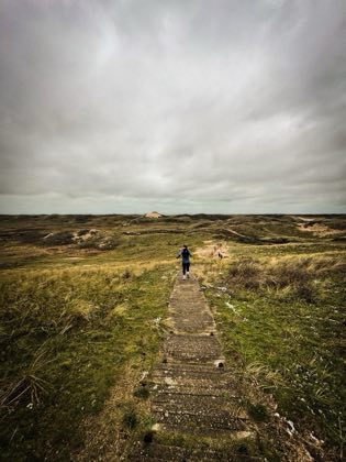 Een houten vlonderpad door de duinen onder een grijze lucht.