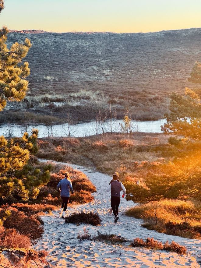 Twee hardlopers op een zandpad door de duinen, met de zon laag achter de heuvels.